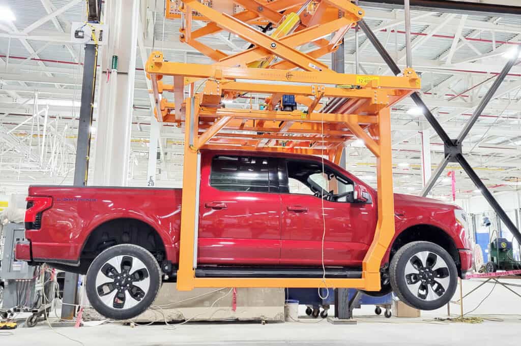 Red truck lifted by orange custom work positioners in an automotive assembly line.