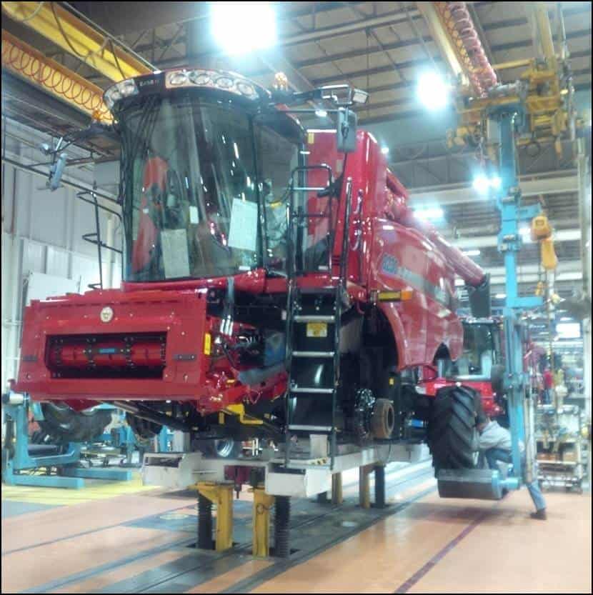 Elevated red agricultural vehicle supported by an industrial hoist system in a manufacturing area.