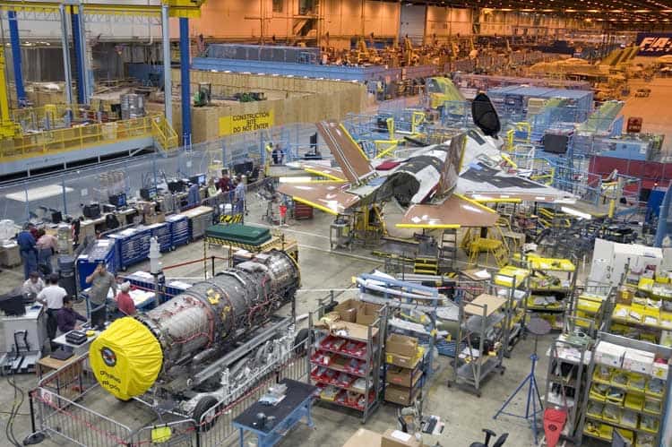 Large industrial facility assembling fighter jets with various workstations, machinery, and aircraft parts.